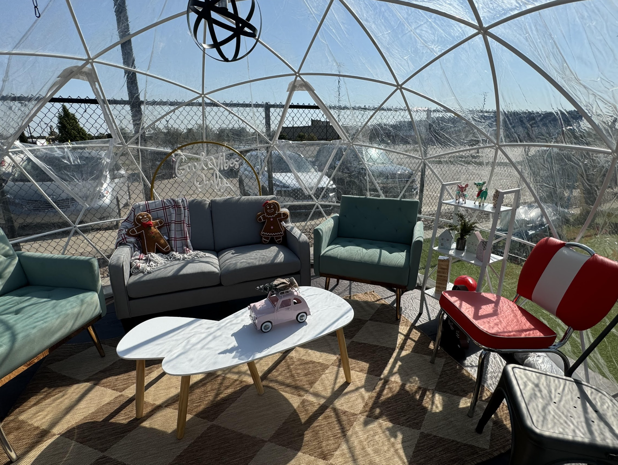 A cozy seating area inside a clear dome features vintage-style chairs, a gray sofa with gingerbread pillows, a white coffee table with a pink toy car, and sunlight streaming through transparent walls.